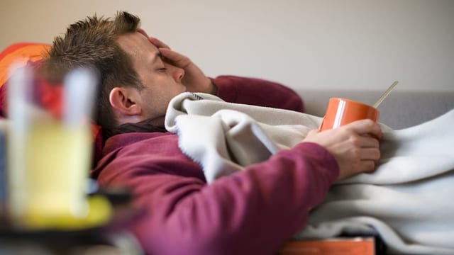 Ein kranker Mann liegt unter einer Decke, hält eine Tasse Tee und fasst sich an den Kopf