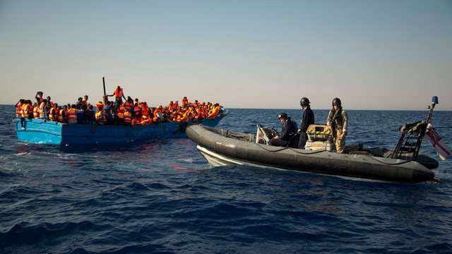 Britisches Schlauchboot nähert sich einem vollbesetzten Holzbötchen im Meer.