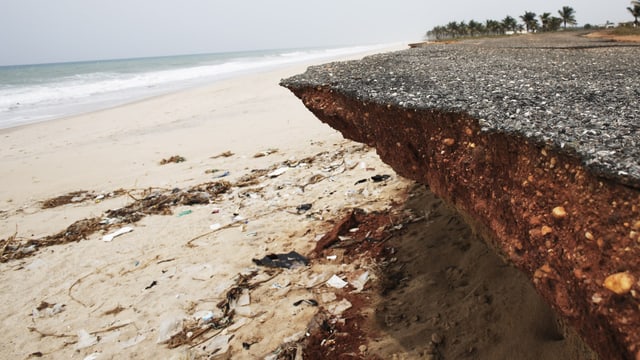 Die an den Rändern abgebrochene und unterspülte Strasse ragt über den Sandstrand.