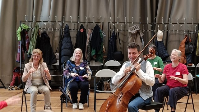 Teilnehmerinnen sitzen auf Stühlen und bewegen sich. In der Mitte sitzt ein Cellist.