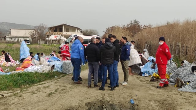 Rettungskräfte, dahinter sitzen Menschen mit dunkler Hautfarbe am Boden. Sie sind in Decken eingehüllt.