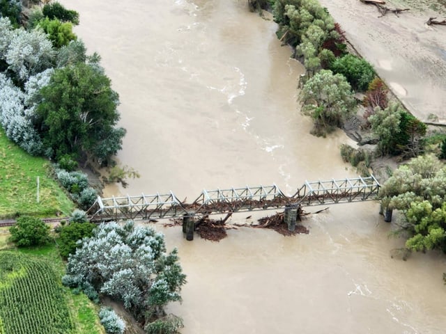 Kaputte braune Brücke steht noch halb über braunem Fluss. Links und rechts vom Fluss sind grüne Bäume zu sehen. 