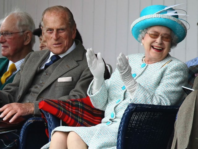 An older man in a brown blazer laughs in front of the camera, next to him an older woman in a blue dress.