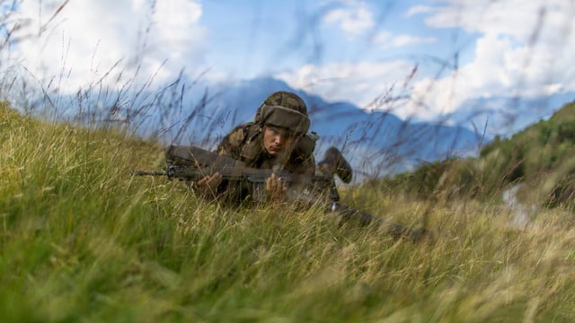 Rekrut liegt mit Waffe im Gras, dahinter Bergpanorama. 