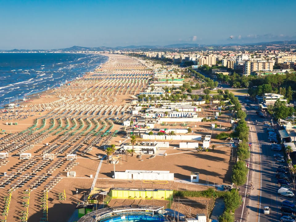 Luftaufnahme eines Strandes mit vielen Liegen und Sonnenschirmen, Stadt im Hintergrund.