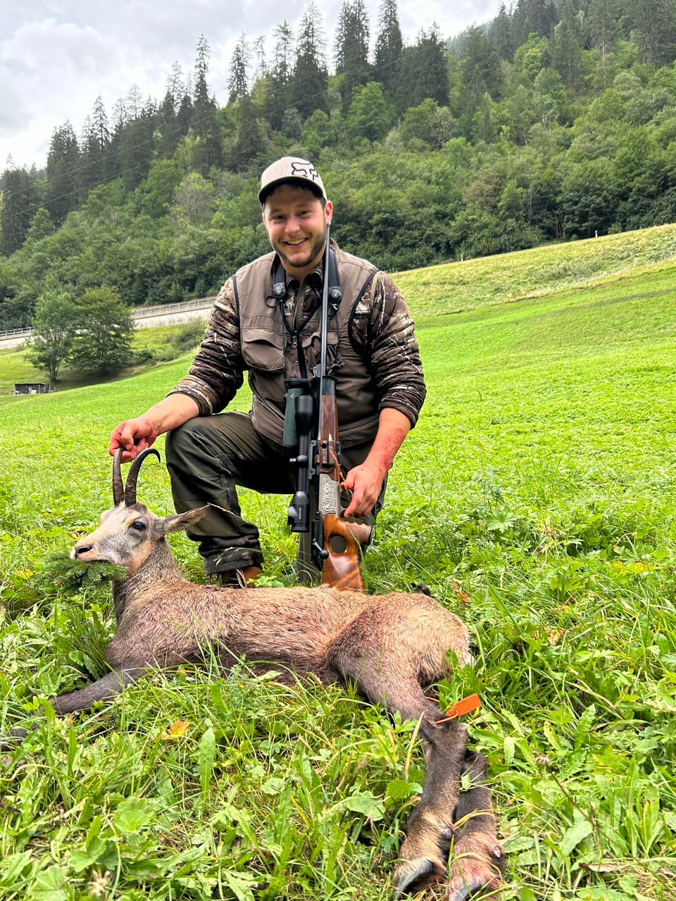 Mann posiert mit erlegtem Steinbock auf einer Wiese vor bewaldetem Hügel.