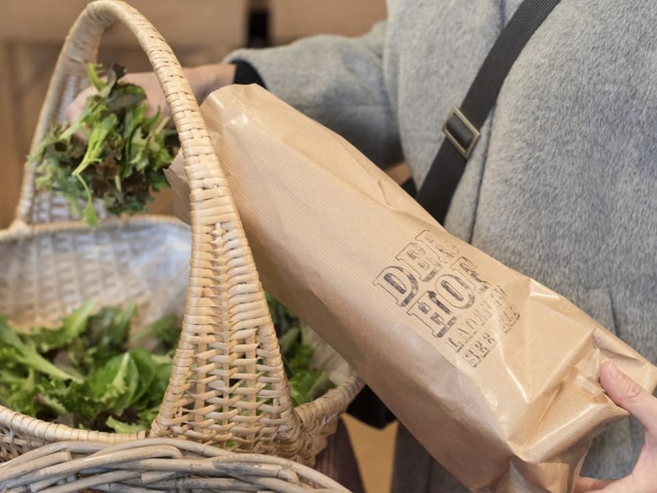 Eine Person hält eine braune Tüte und greift in einen Korb mit Salat.