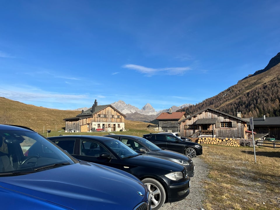 Parkplatz mit Autos vor Berghütten und Bergen im Hintergrund.