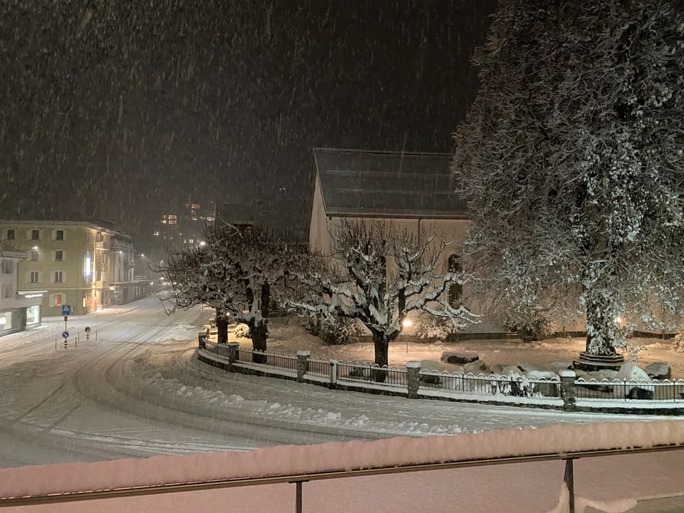 Schneebedeckter Platz mit Bäumen und Häusern bei Nacht.