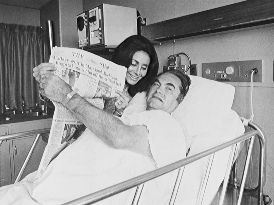 Mann im Krankenhausbett liest Zeitung, Frau lächelt neben ihm.