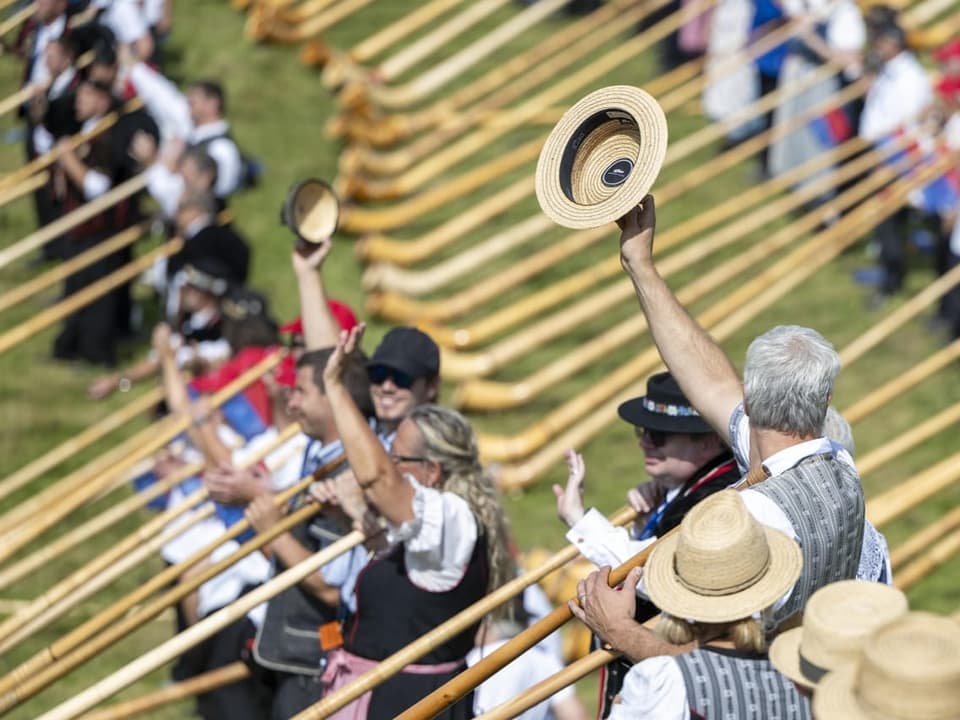 The horn players hold their alphorn with one hand and lift parts of their hut with the other.