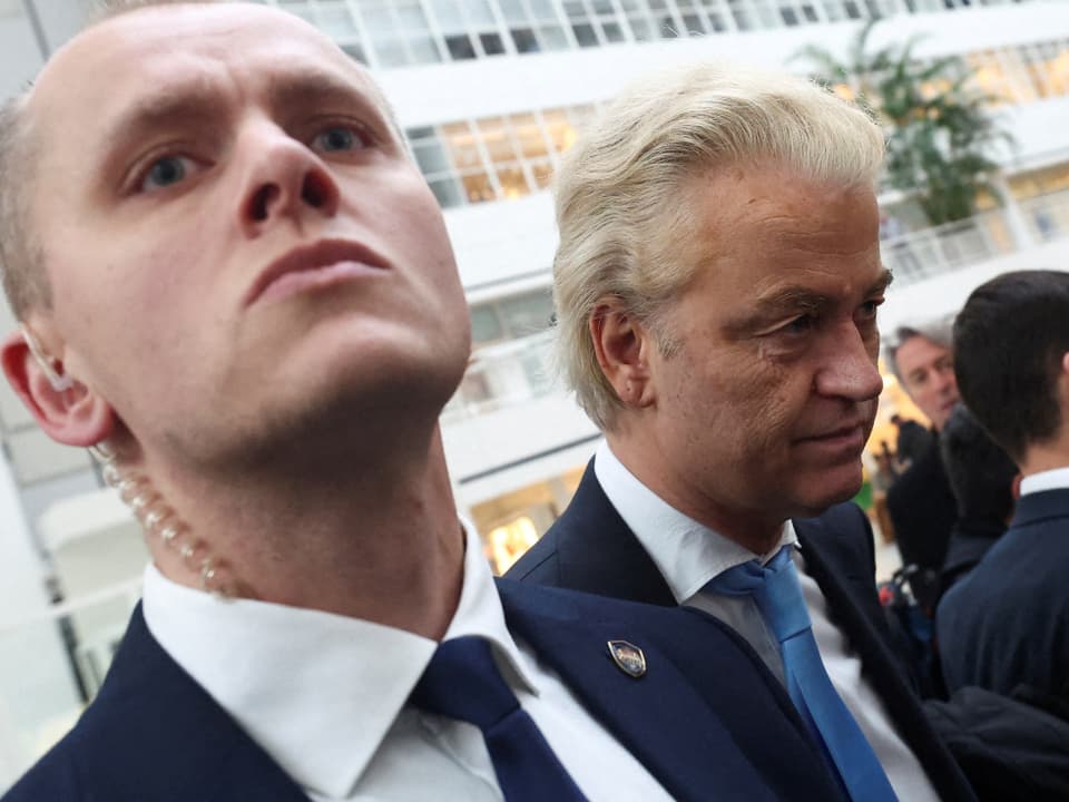 Ein Sicherheitsguard schaut angespannt über die Kamera. Er steht direkt neben Geert Wilders, der auf den Boden blickt.