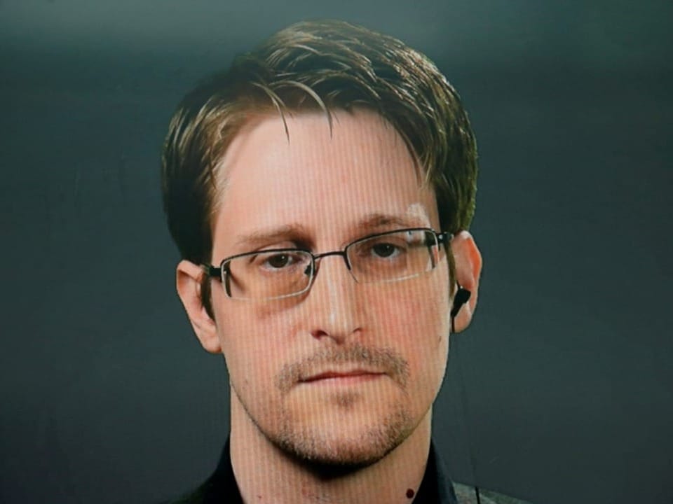 Porträt eines Mannes mit Brille und Ohrhörer vor grauem Hintergrund.