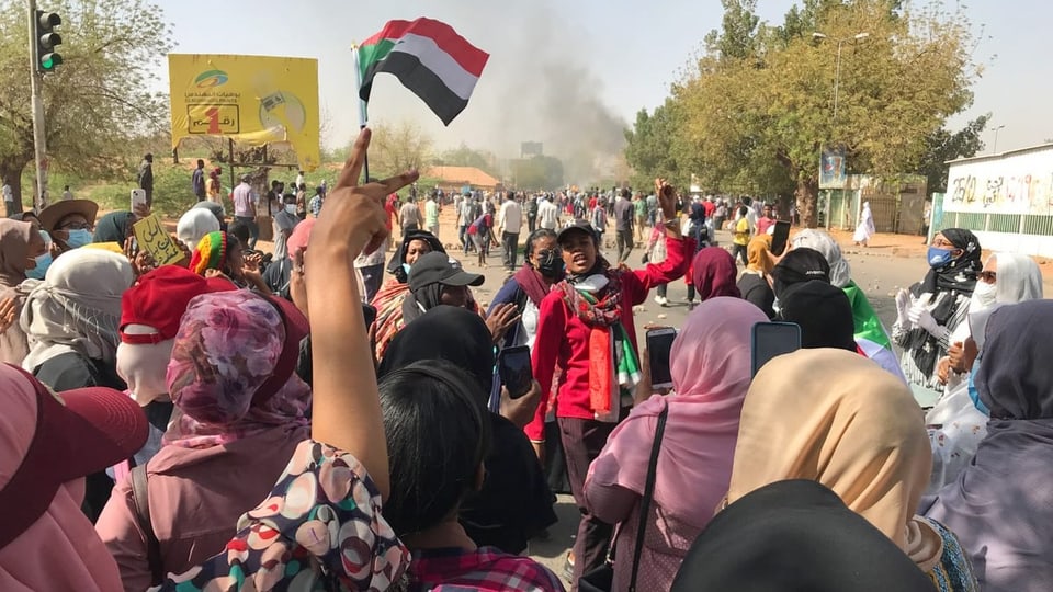 Viele Frauen stehen auf einer Strasse und demonstrieren mit einer Flagge.
