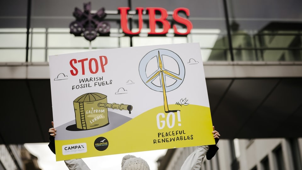 Person hält Protestplakat vor UBS-Gebäude mit Aufschrift 'Stop Warring Fossil Fuels, Go Peaceful Renewables'.