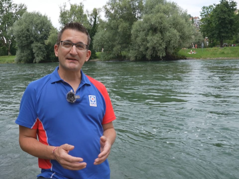 Mann in blauem Hemd steht vor einem Fluss mit Bäumen im Hintergrund.