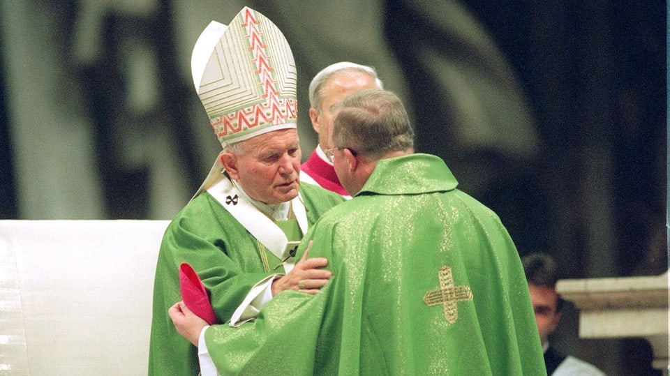 Ein alter Mann mit Mitra auf dem Kopf und grünem Priestergewand umarmt einen anderen älteren Mann, ebenfalls in Grün.