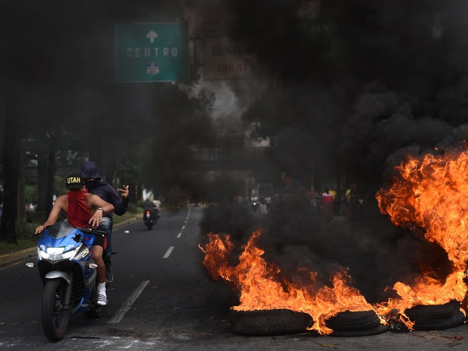 Reifen auf einer Strasse stehen in Flammen. Zwei vermummte Personen fahren mit einem Motorrad neben den Reifen durch.