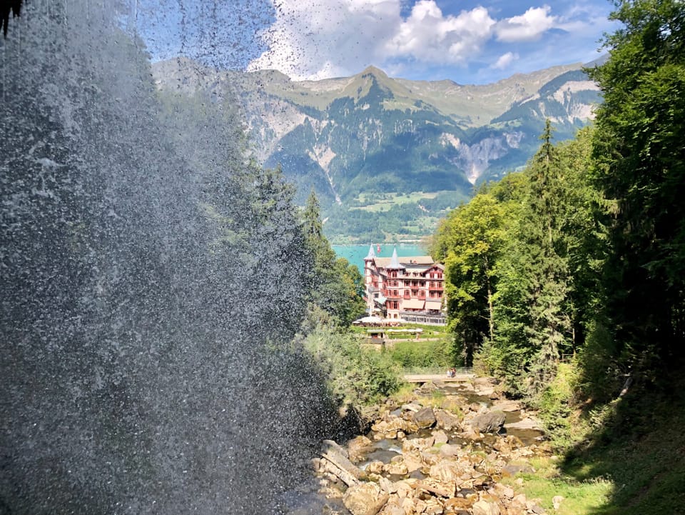 Man sieht die Tropfen des Wasserfalls auf der Seite, während der Blick zum Hotel und dem dahinterliegenden See schweift.