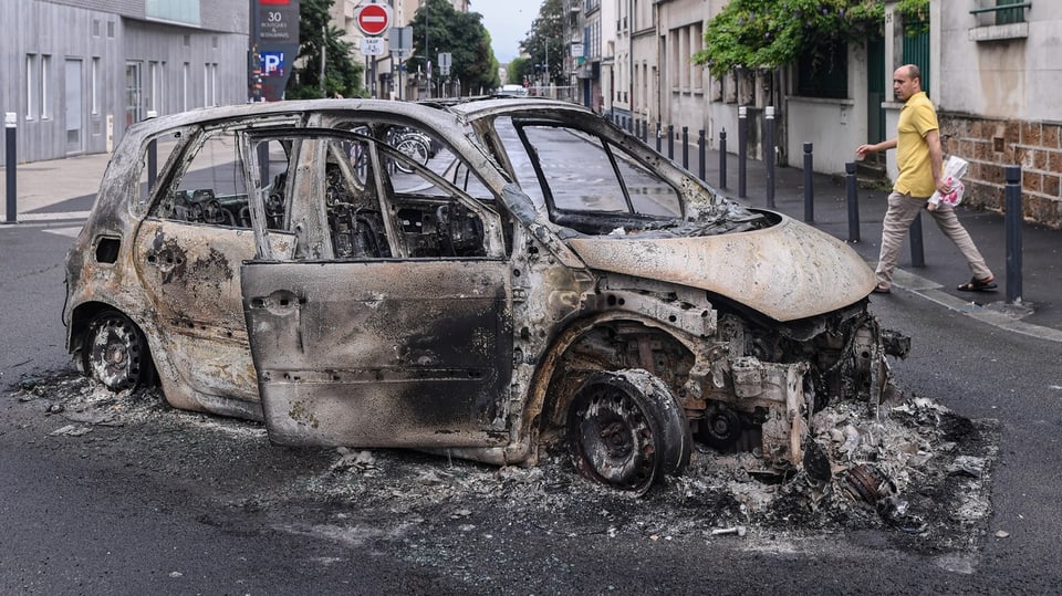 Ein komplett abgebranntes Auto steht auf einer Strasse mitten am Tag.