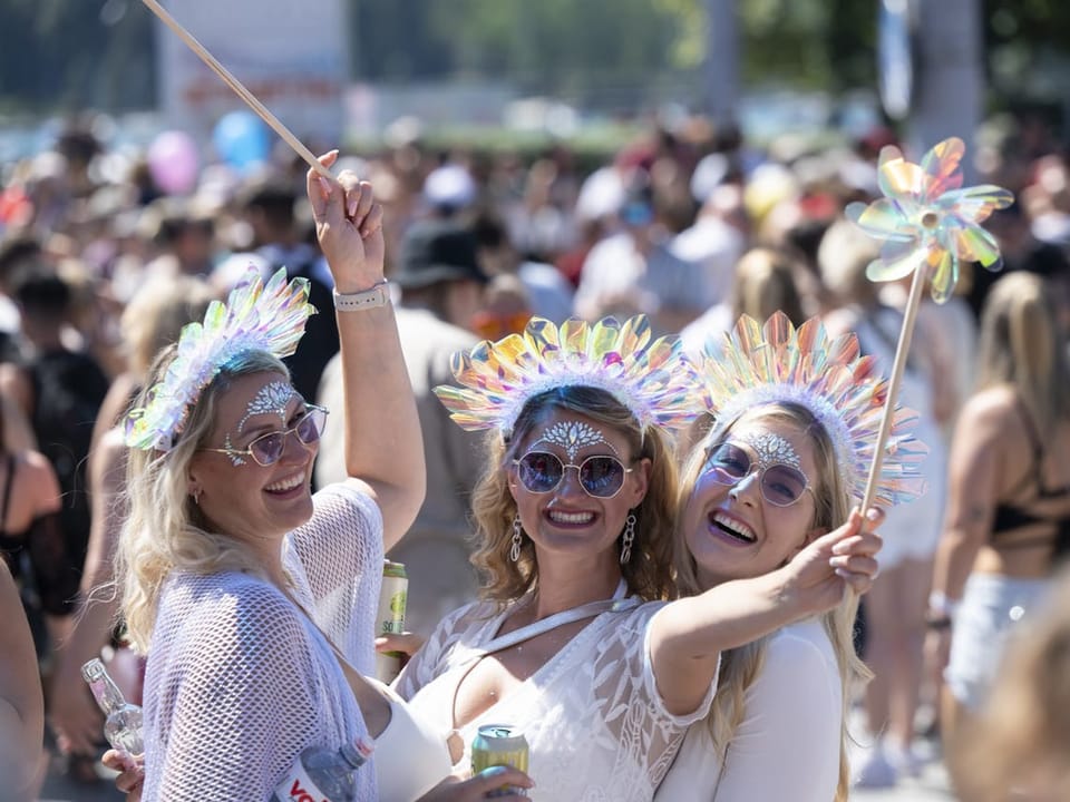 Drei Frauen mit bunten Hüten und Brillen, die bei einer Menschenmenge feiern.