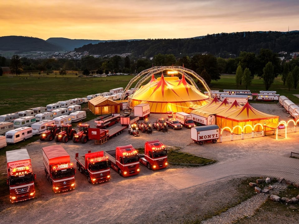 Luftaufnahme eines beleuchteten Zirkuszeltes mit roten Lastwagen bei Sonnenuntergang.