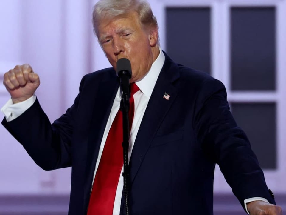 Mann in dunkelblauem Anzug und roter Krawatte hält eine Rede.