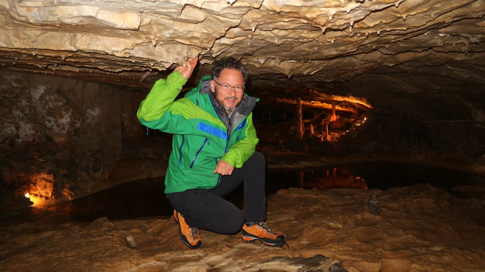 Ein Mann kniet in Wanderausrüstung in einer Höhle und grinst.