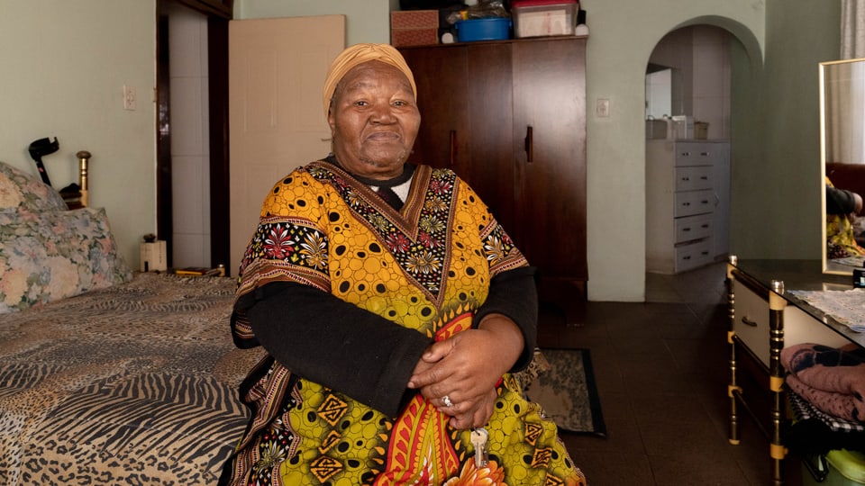 Selina Mhabela sitzt auf einem Bett mit Leoparden- und Zebramuster. Sie trägt ein langes Kleid mit Afrikamuster. 