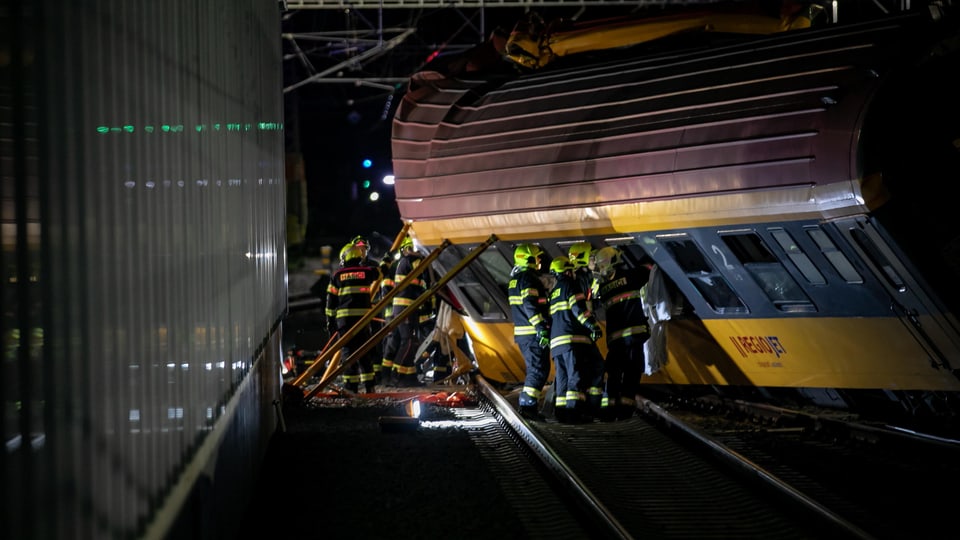 Nachtszene eines entgleisten Zugs mit Feuerwehrleuten bei der Arbeit.