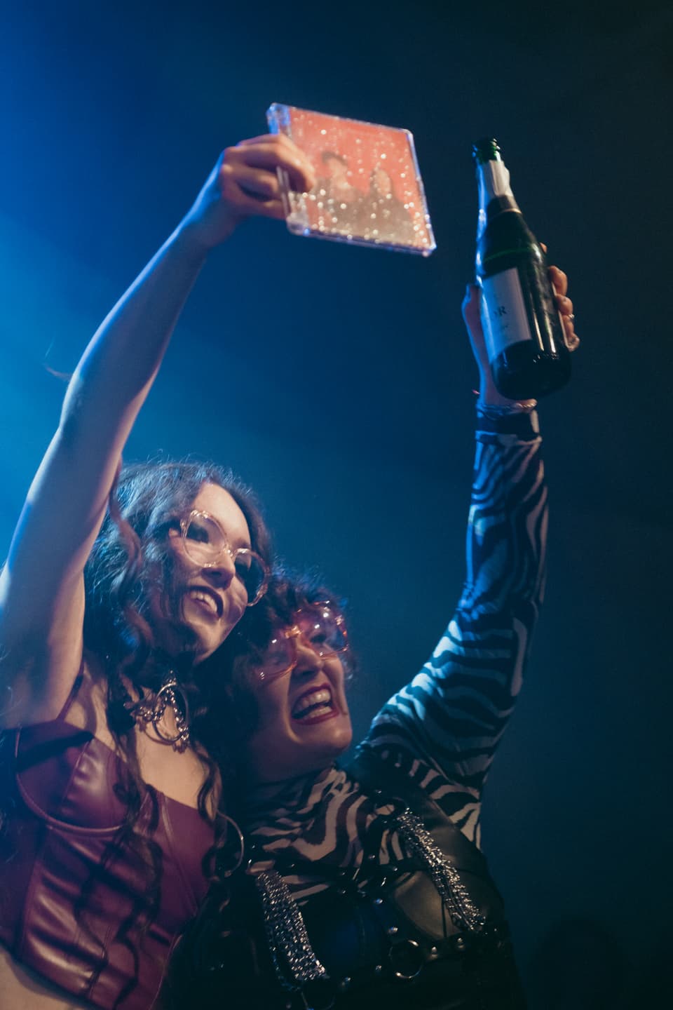 Chiara Jacomet und Momo Kawazoe lachen. Momo hebt eine CD in eine Hand und Chiara eine Champagnerflasche in einer Hand