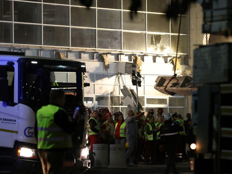 Bauarbeiter in Schutzwesten vor einem beleuchteten Gebäude bei Nacht.