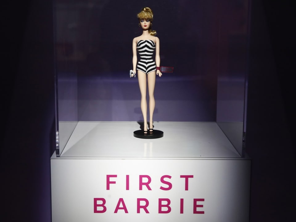 Die erste Barbie trägt einen schwarz-weiss gestreiften Einteiler und steht ausgestellt in einem Glaskasten. 