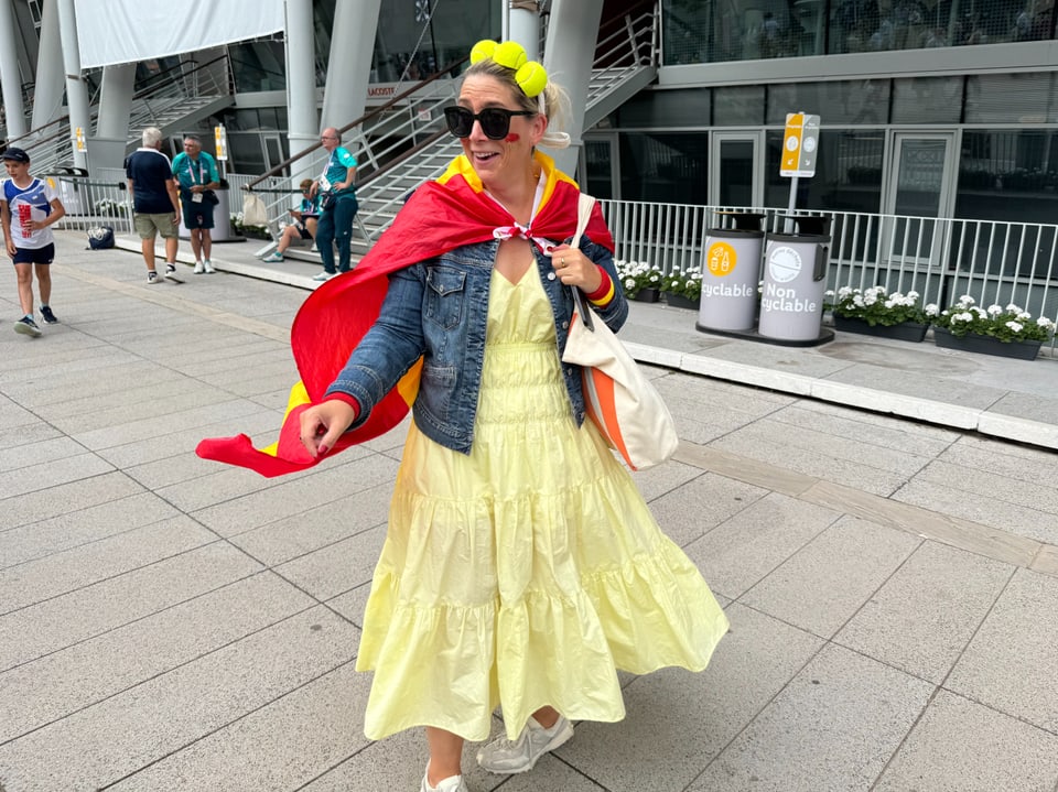 Frau mit Sonnenbrille, gelbem Kleid und rotem Cape vor einem Gebäude.