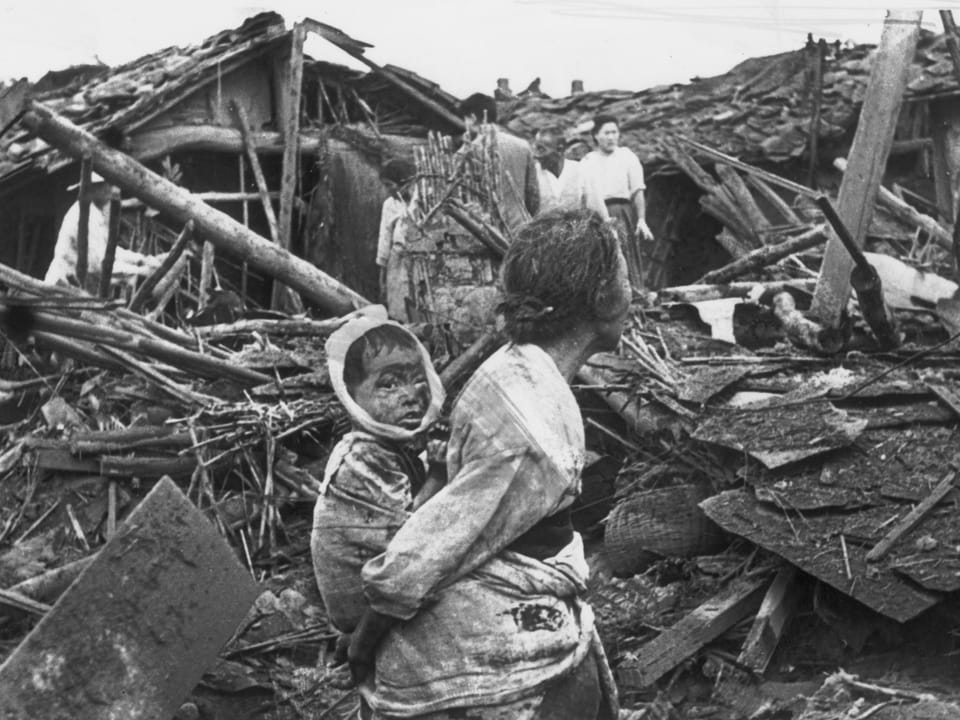 Frau mit Kind im Tragetuch vor zerstörten Häusern.