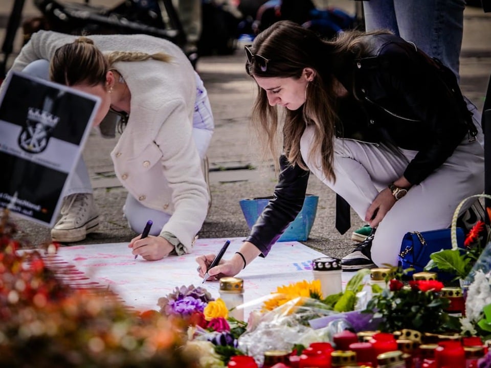 Zwei Frauen schreiben auf ein Banner bei einer Gedenkstätte mit Blumen.