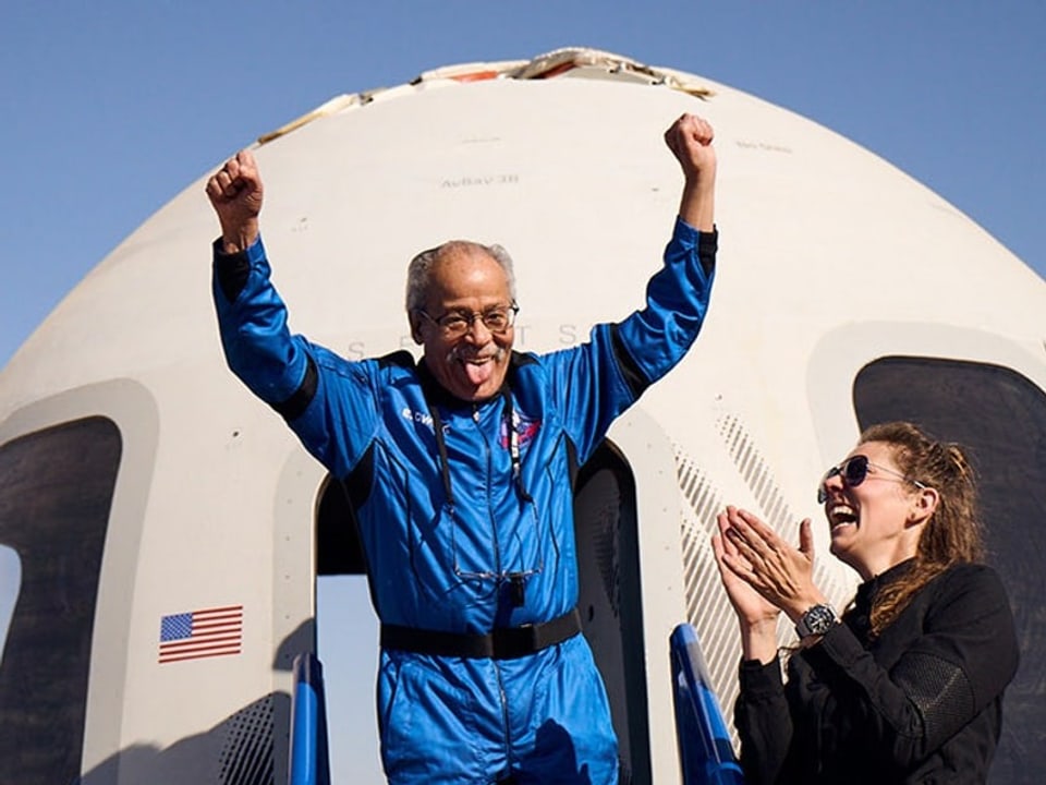 Mann in blauer Raumanzug jubelt neben einer Frau vor einer Raumkapsel.