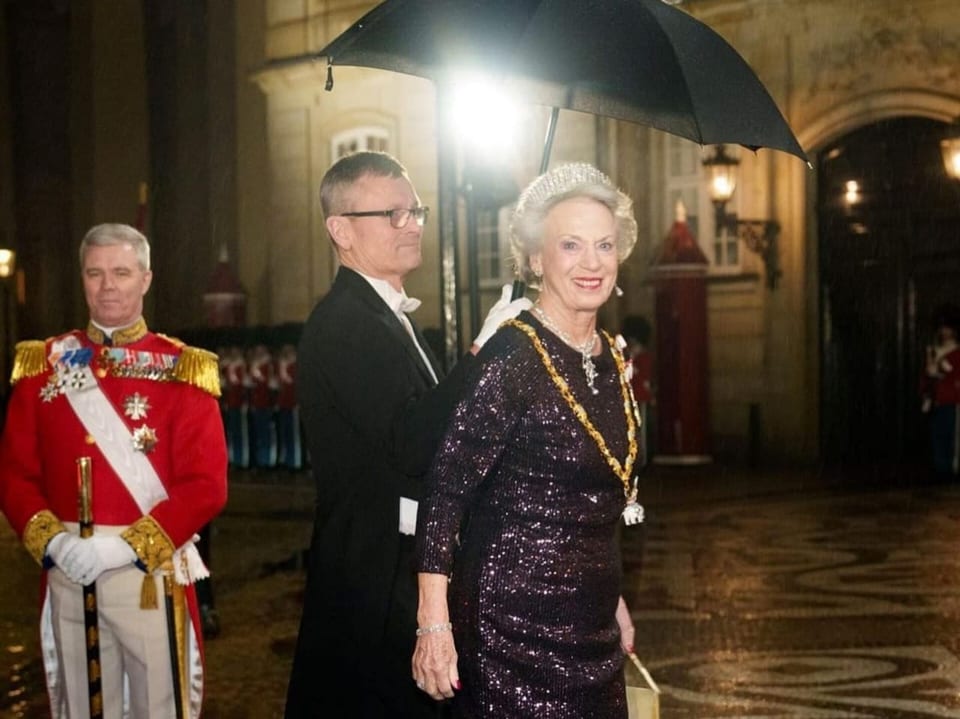 Eine ältere Dame in einem edlen glitzernden Kleid wird von einem Mann mit Regenschirm begleitet. 