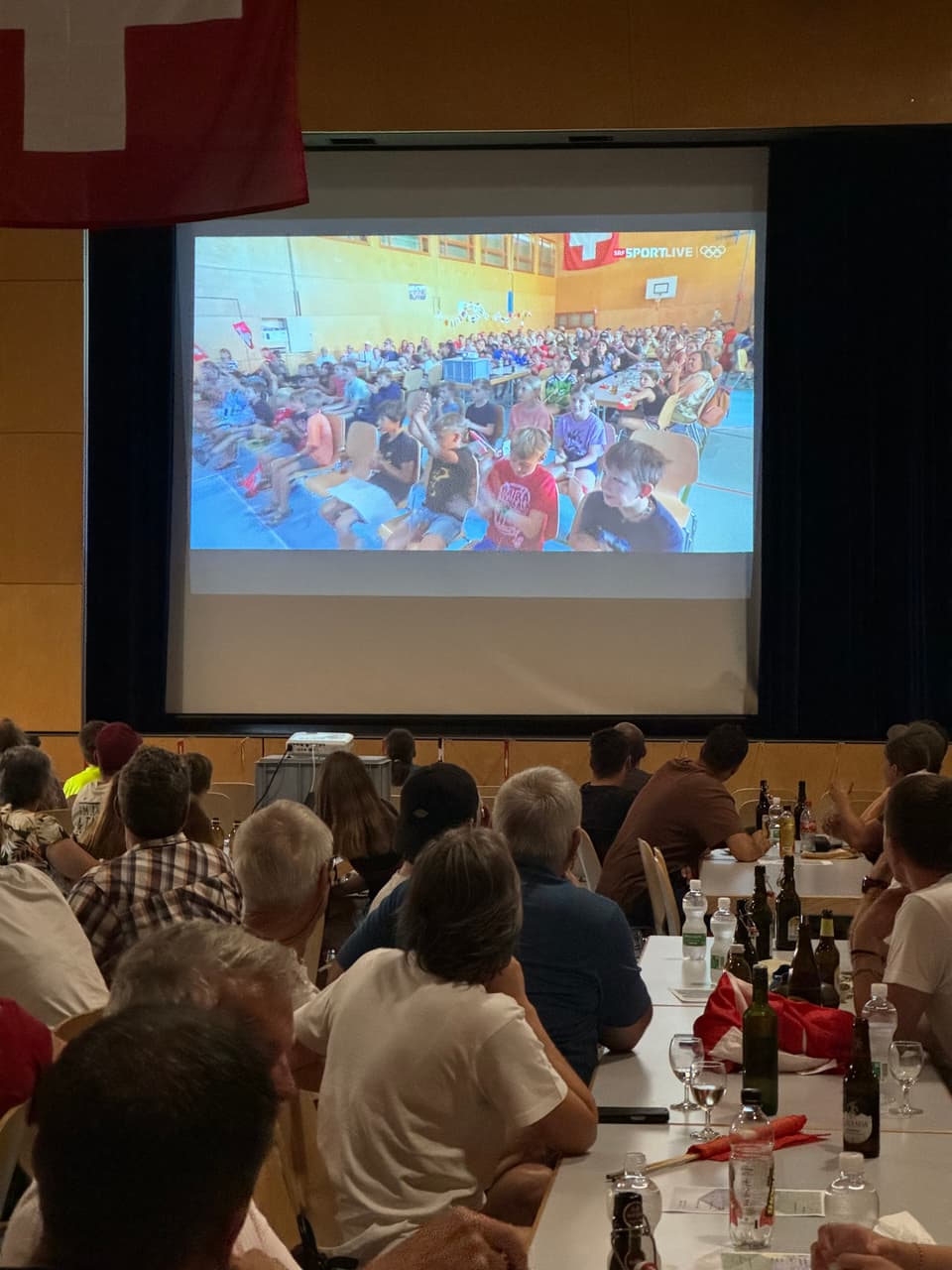 Personengruppe verfolgt einen Sportevent auf einer Leinwand in einer Halle.