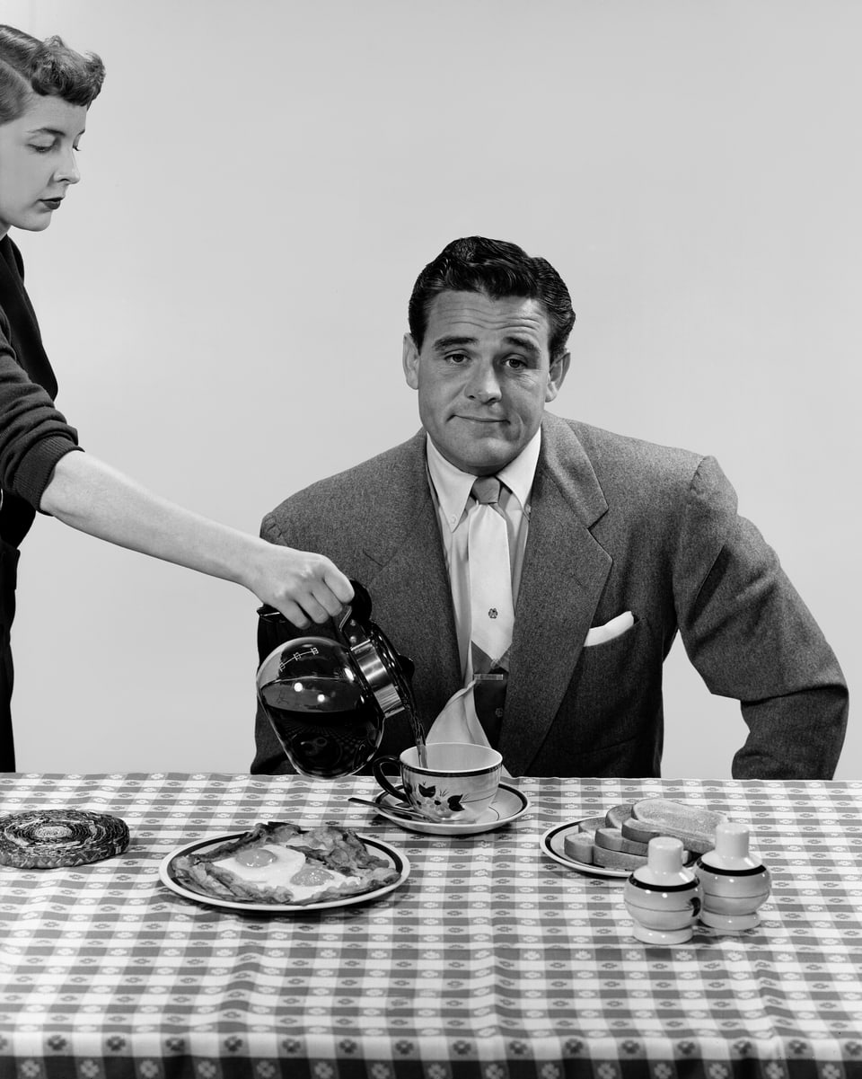 Ein Mann guckt unbegeistert in die Kamera vor seinem Frühstück. Die Frau schenkt ihm Kaffee ein.
