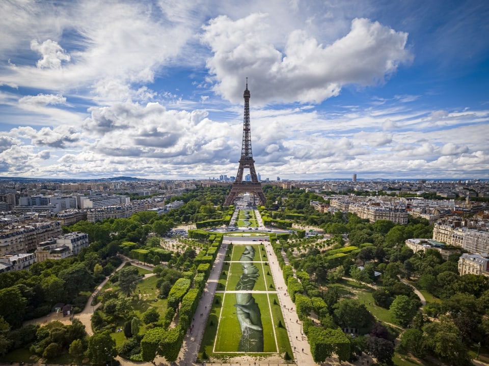 Ein Bodenbild, das Arme zeigt, ineinander verschlungen. Das Bild ist aufgemalt auf eine Rasenfläche vor dem Eiffelturm.