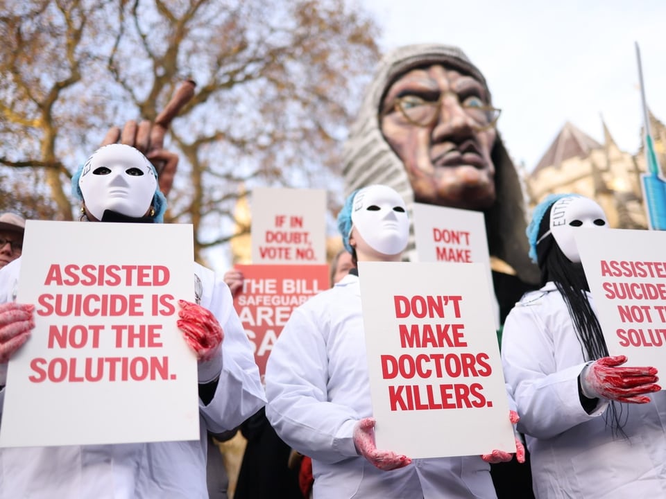 Protestierende mit Masken halten Schilder gegen Sterbehilfe.