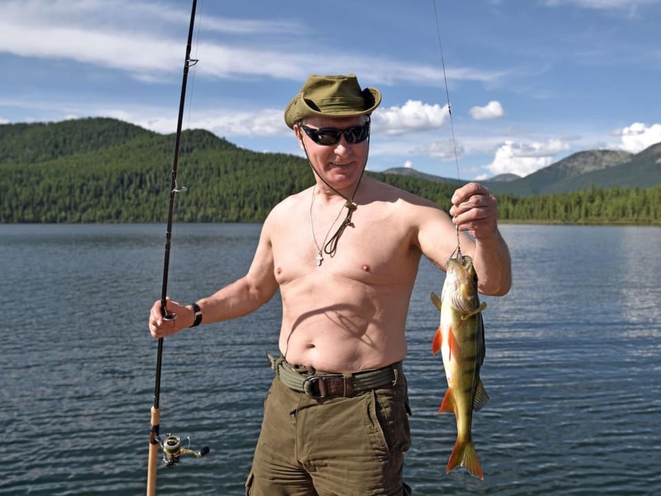 Putin hält einen Fisch in der Hand und steht mit nacktem Oberkörper und Sonnenhut am Ufer eines Sees.