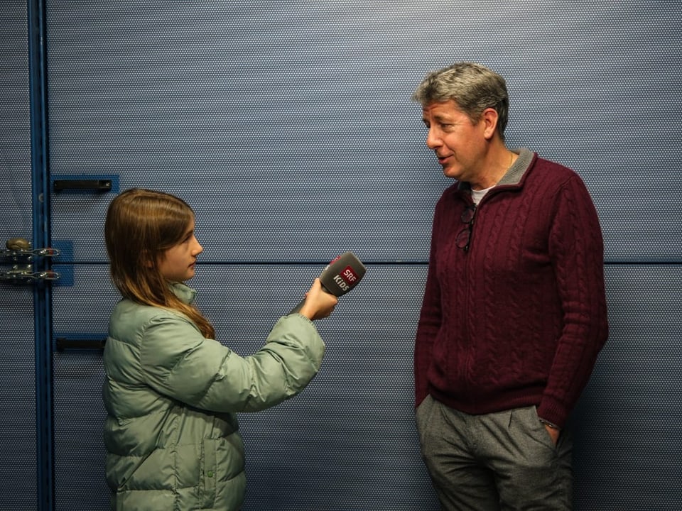 Ein Mädchen interviewt einen Mann. Hinter ihr ist eine blaue Wand zu sehen. 