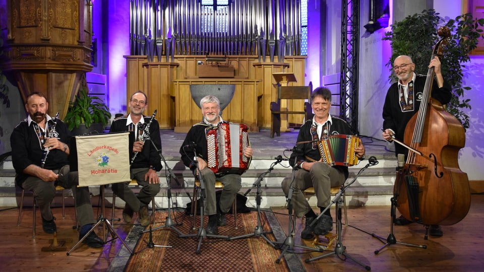 Ländlerkapelle Hoharätia, zu fünft mit den Instrumenten, sitzend, alle in dunkelgrauen Hosen und schwarzer Sennenkutte.