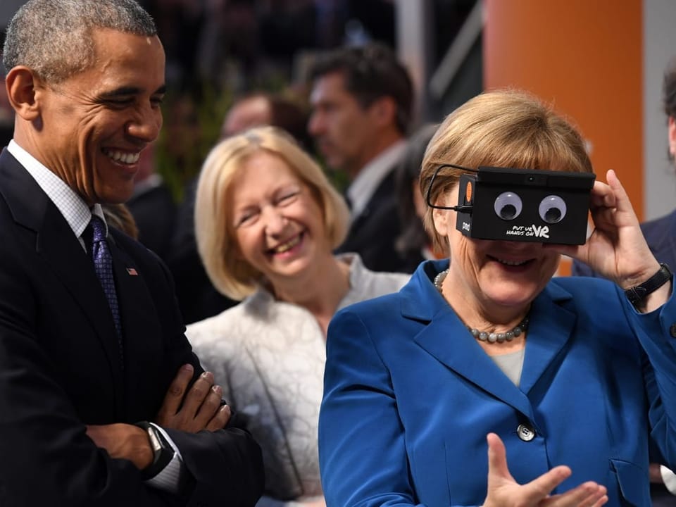Angela Merkel hält sich ein Spielzeug vors Gesicht, eine schwarze box mit zwei Augen