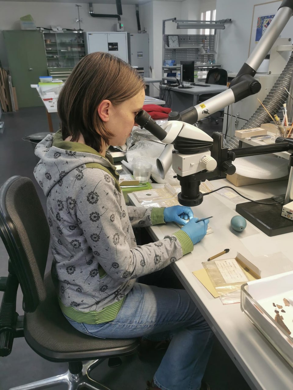 Johanna schaut durch ein Binokular auf eine archäologische Münze und hält ein Skalpell in der Hand. 