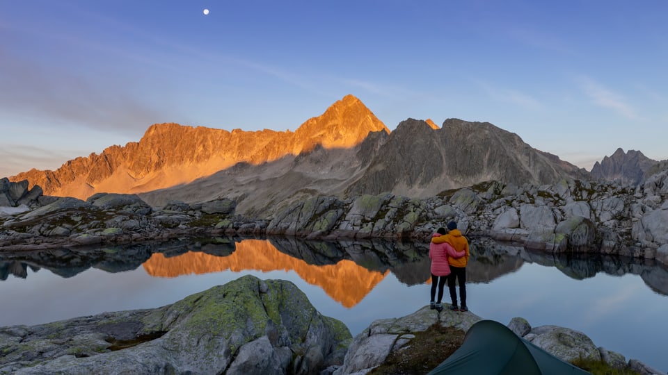 Zwei Personen vor alpenländischer Berglandschaft mit See bei Sonnenaufgang.