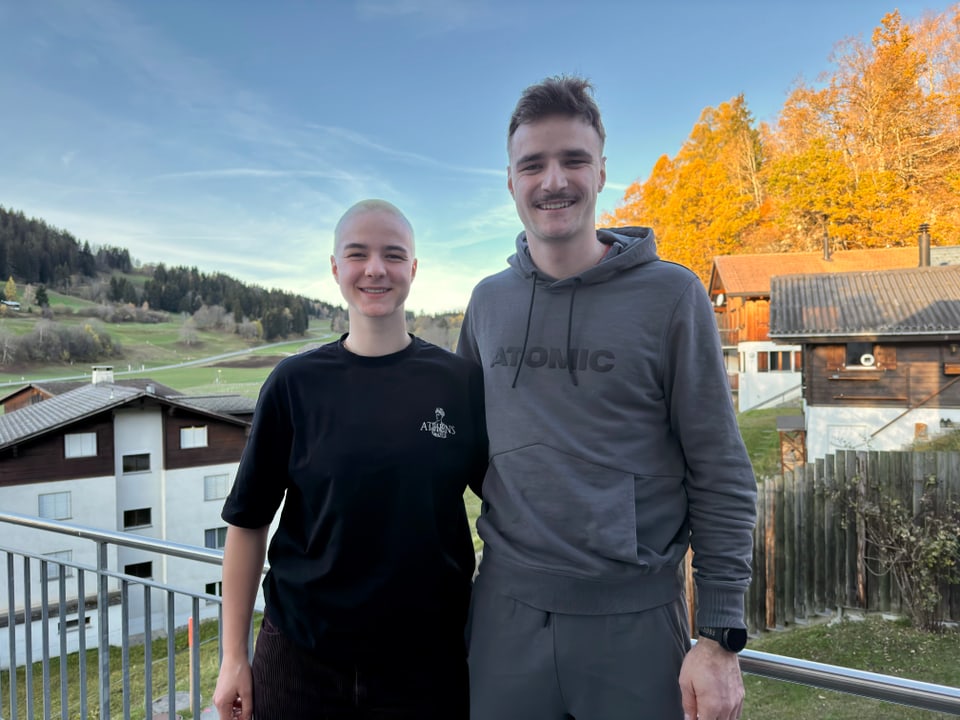 Zwei Personen lächeln auf einem Balkon mit Herbstlandschaft im Hintergrund.