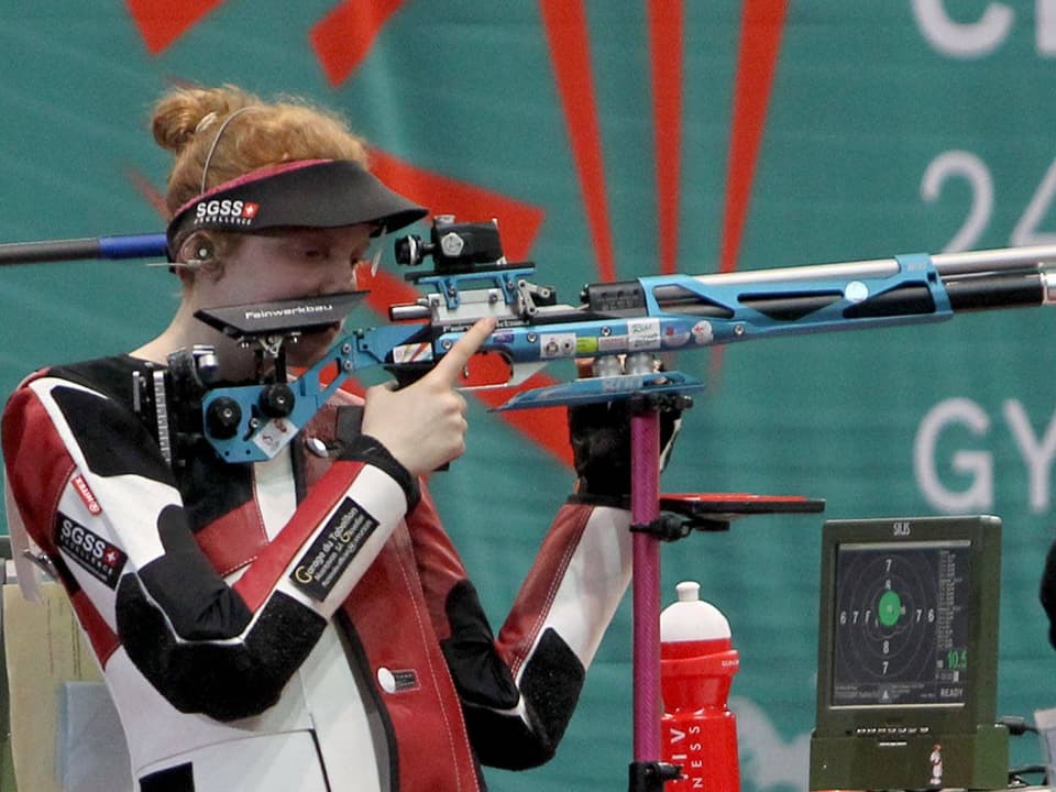 Schütze in Sportschiesskleidung zielt mit Gewehr auf Zielscheibe.